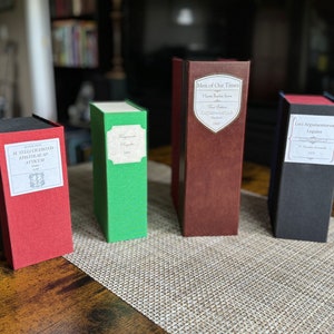 Puede incluir: Cuatro cajas decorativas con forma de libro en rojo, verde, marrón y negro. Cada caja tiene una etiqueta blanca con texto. Las cajas están dispuestas sobre una mesa con un mantel individual tejido de color neutro.