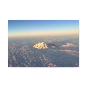 May include: An aerial view of a snow-capped mountain peak rising above a sea of clouds. The mountain is illuminated by sunlight, with a clear blue sky above. The clouds are a mix of white and gray, creating a dramatic landscape.