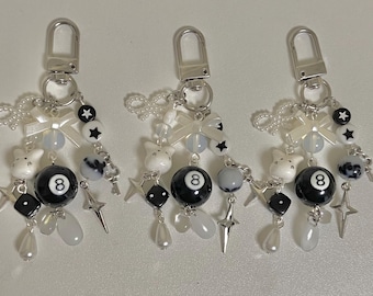 May include: Three black and white keychains with a silver charm. Each keychain features a white cat charm, a black 8-ball charm, a white star charm, a white pearl charm, a silver star charm, and a silver dice charm.