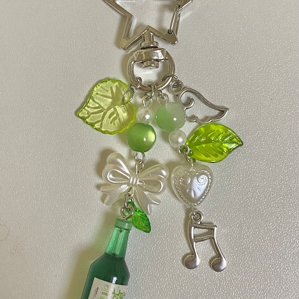 May include: A green and white charm keychain with a miniature bottle of soju, a bow, a heart, a music note, and green leaves. The keychain is attached to a silver star-shaped clip.