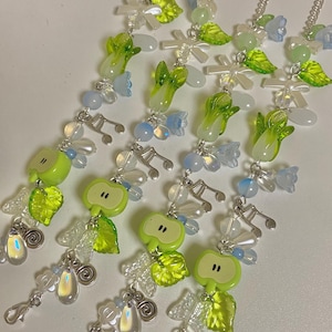 Könnte beinhalten: Eine Sammlung skurriler Charms mit grünen Apfelscheiben, Blättern und Noten. Die Charms sind mit klaren und irisierenden Perlen sowie kleinen blauen Blumenakzenten verziert, die alle an silbernen Ketten hängen.