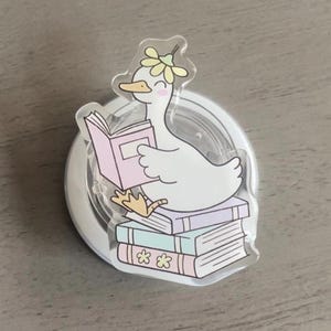 May include: A clear acrylic charm featuring a white cartoon goose wearing a flower crown and reading a pink book while sitting on a stack of pastel-colored books. The charm is on a clear circular base.
