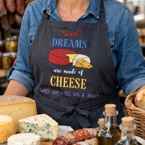 Könnte beinhalten: Eine schwarze Schürze mit gesticktem Text Sweet DREAMS are made of CHEESE WHO AM I TO DIS A BRIE? und einer bunten Grafik, die einen roten Käselaib, ein Stück weißen Käse und ein Stück gelben Käse in der Mitte zeigt.