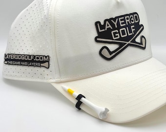 May include: White golf hat with the text "LAYER3D GOLF" and a logo of crossed golf clubs. The hat has a perforated side panel and a golf tee holder with a white and yellow tee. The text "LAYER3DGOLF.COM" is also visible.