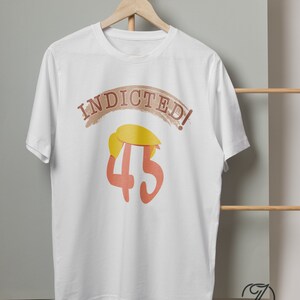 May include: White t-shirt with the text "INDICTED!" in a brown arch above the number "45" in orange. The number has a yellow cartoon hair style on top.