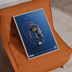 Peut inclure: Affiche encadrée représentant un joueur de football en uniforme bleu et jaune, sur fond bleu foncé avec le logo Boca Juniors. L'affiche est posée sur un fauteuil orange. Le nom du joueur est Martin Palermo.