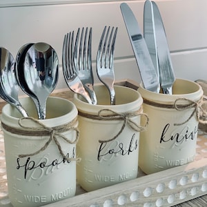 May include: Three white painted mason jars with twine tied around the top, labeled "Spoons", "Forks", and "Knives".  The jars are filled with silverware, including spoons, forks, and knives.