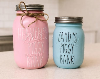 May include: Two painted mason jar piggy banks. One is pink with "Heaven's Piggy Bank" in white script, tied with twine. The other is light blue with "Zayd's Piggy Bank" in black lettering. Both have silver lids.