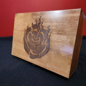 May include: A wooden box with a dark brown rose and flame design. The box is rectangular and made of light brown wood. The rose design is etched into the wood, creating a contrasting dark brown color. The box is on a black surface.