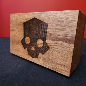 May include: A wooden block with a skull design. The skull is dark brown and geometric, with two circular eye sockets and a small nose. The block is made of light brown wood with visible grain.