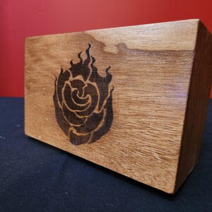 May include: A wooden box with a dark rose design burned into the wood. The box is rectangular and has a natural wood grain finish. The rose design is centered on the front of the box.