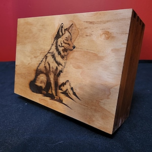 May include: Wooden box with a detailed pyrography image of a coyote. The artwork is burned into the light-colored wood, showcasing the animal's fur and features. The box is rectangular and has a natural wood finish.