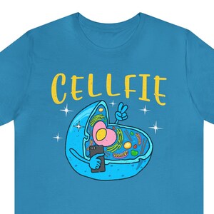 May include: A turquoise t-shirt with a cartoon cell design. The cell, in blue, is taking a selfie with a smartphone. The word "CELLFIE" is written in yellow above the cell, with a peace sign. The cell has a pink nucleus.