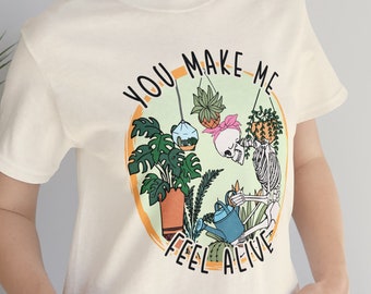 May include: Cream-colored t-shirt featuring a whimsical design with a skeleton tending to plants. The graphic includes the text "YOU MAKE ME FEEL ALIVE" in a circular design with orange and green accents. The shirt is made of soft material.