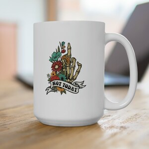 Peut inclure: Mug en céramique blanche avec une main de squelette ornée de fleurs et l'inscription "NOT TODAY" sur une bannière. Le motif floral comprend des fleurs turquoise, orange et jaunes. La tasse a une grande anse et est posée sur une surface en bois.