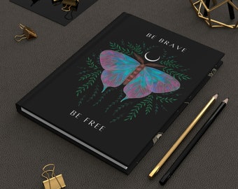 May include: Black journal with a colorful butterfly design and the words "BE BRAVE BE FREE". The butterfly has blue, purple, and teal wings. Gold and black pencils and gold binder clips are also visible.