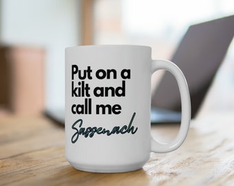 May include: White ceramic mug with the text "Put on a kilt and call me Sassenach" in black and teal lettering. The mug has a large handle and is sitting on a wooden surface.