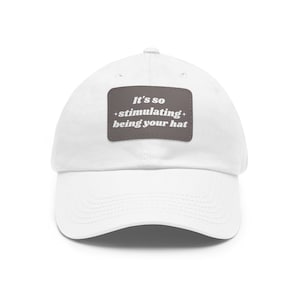 May include: White baseball cap with a curved brim. A rectangular patch on the front displays the text "It's so + stimulating + being your hat" in white letters on a gray background. The hat is made of a soft fabric.