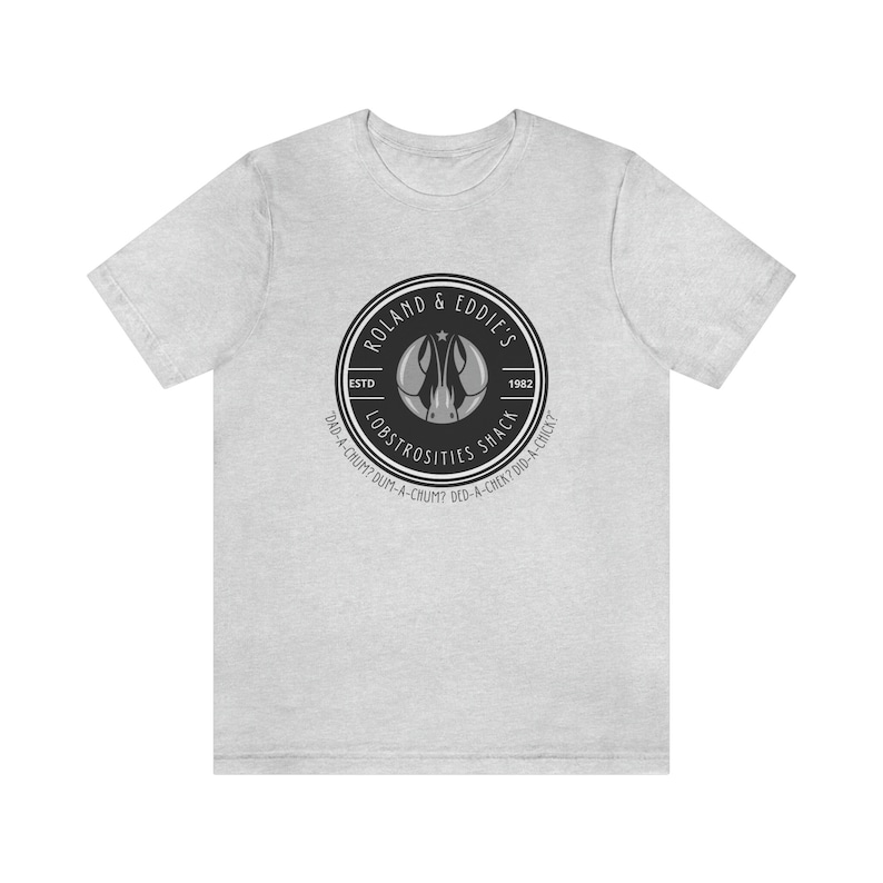 Puede incluir: Una camiseta gris con un logotipo en blanco y negro para "Roland & Eddie's Lobstrosities Shack" establecido en 1982. El logotipo presenta un langostino con una estrella en la cabeza.