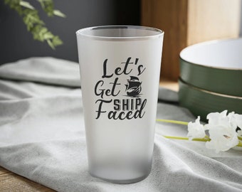 May include: A frosted glass with the phrase "Let's Get Ship Faced" in black script and a ship illustration. The glass is cylindrical and sits on a light gray cloth. The background includes a dark gray wall and a wooden surface.