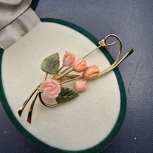 Puede incluir: Broche dorado con diseño floral. El broche presenta flores de coral rosa y hojas verdes, sobre un tallo dorado. El broche se presenta en un joyero de terciopelo verde.
