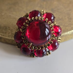 Peut inclure: Broche vintage avec un groupe de pierres rondes en verre rouge foncé. La pierre centrale est plus grande, entourée de pierres plus petites et d'une monture en filigrane dorée. La broche a un design classique et élégant.