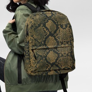 Può includere: Uno zaino con motivo a pelle di serpente nei toni del marrone, beige e nero. Lo zaino ha una tasca frontale e spallacci neri. La borsa è indossata da una persona che indossa una giacca verde e jeans neri.