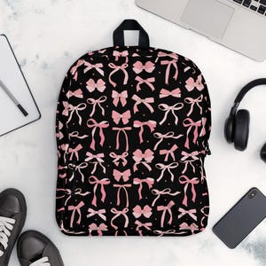 Könnte beinhalten: Schwarzer Rucksack mit einem Muster aus rosa Schleifen. Der Rucksack hat einen Reißverschluss und verstellbare Träger.