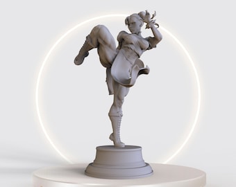 May include: A gray 3D printed figurine of a female character in a fighting pose, standing on a gray base. The base is on a white platform. The text "STL FALLEN" is printed in gold letters on the platform.