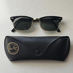 May include: Black and gold browline sunglasses with dark lenses, featuring a classic frame design. Accompanied by a black leather-like protective case with a circular gold emblem printed with the text 100% UV protection Ray-Ban Bausch & Lomb.