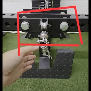 May include: A black weight machine attachment with a metal frame, bolts, and a chain. The machine is designed for weightlifting and is set against a backdrop of weight plates and a green floor.