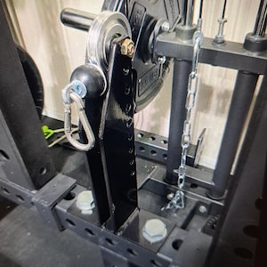 Puede incluir: Una máquina de levantamiento de pesas de metal negro con un sistema de poleas y una cadena. La polea está unida a un marco de metal negro con un mosquetón.