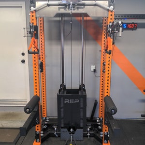 May include: A weight training machine with a black and orange color scheme. The machine has a REP logo and includes various attachments, such as handles and footrests. The frame is orange, and the weight stack is black.