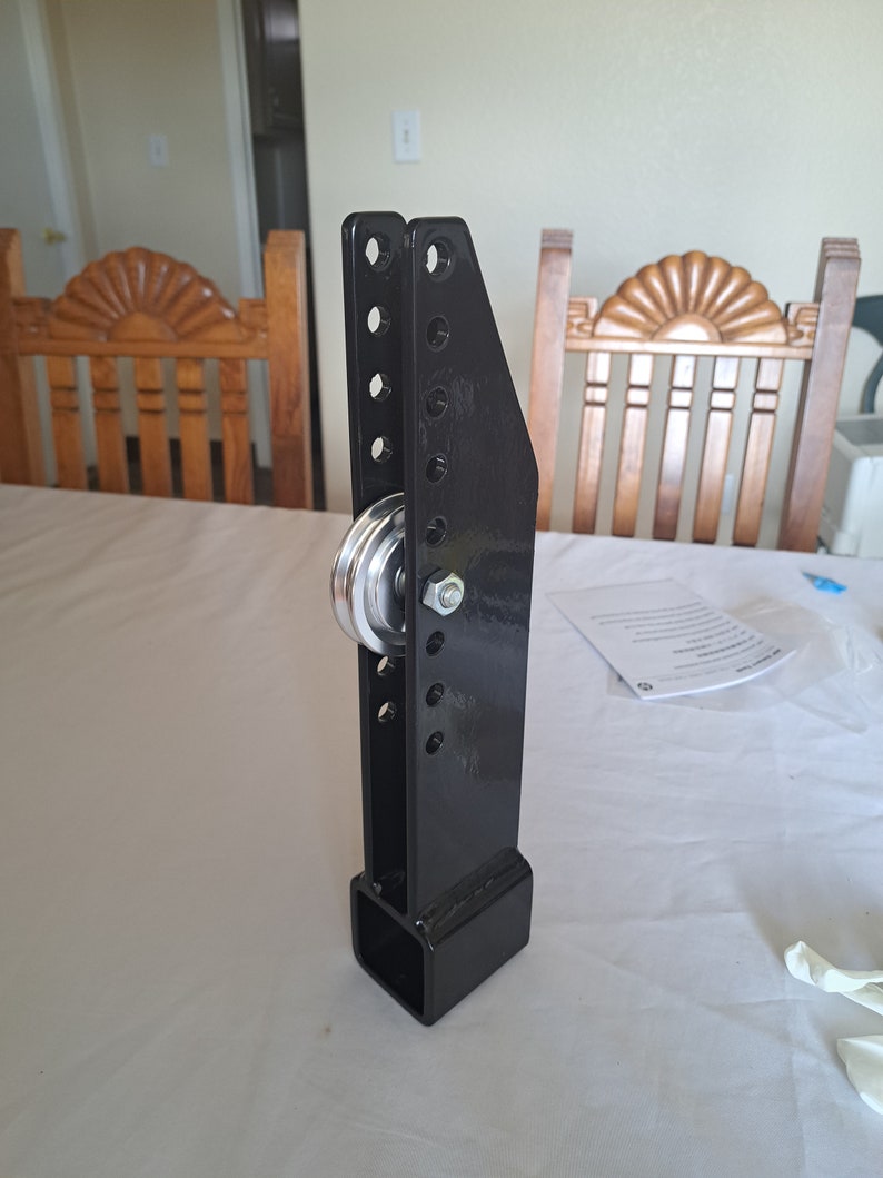 May include: A black metal pulley system with a silver pulley wheel and bolt. The system has a rectangular base and a vertical support with multiple holes. The pulley is designed for lifting or moving heavy objects.