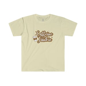 May include: A cream-coloured t-shirt featuring the words "Caffeine Junkie" in brown script, alongside a coffee cup graphic. The shirt has a crew neck and short sleeves.