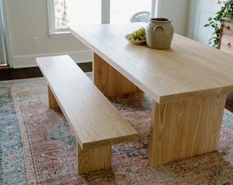 May include: A light wood dining table with a matching bench. The table has a simple, modern design with a rectangular top and a single, thick leg on each side. The bench is also rectangular with a single, thick leg on each end. The table and bench are both made of light wood with a natural finish. The table and bench are set on a patterned rug.