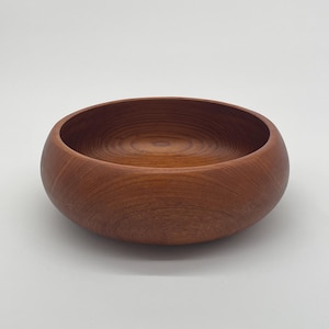 May include: A round wooden bowl with a reddish-brown color. The bowl is made of wood with a smooth, polished finish.
