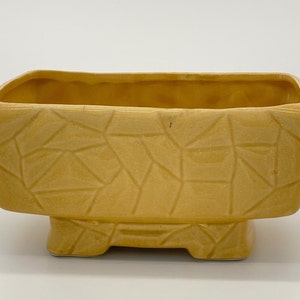 May include: A yellow ceramic planter with a geometric pattern and four short legs.