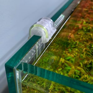 May include: A clear plastic aquarium light fixture with a white clip on the edge of a glass aquarium. The aquarium is filled with green and red aquatic plants.