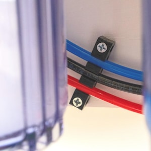 May include: Close-up of three black plastic hose clamps securing three different colored hoses to a white surface. The hoses are red, blue, and black.