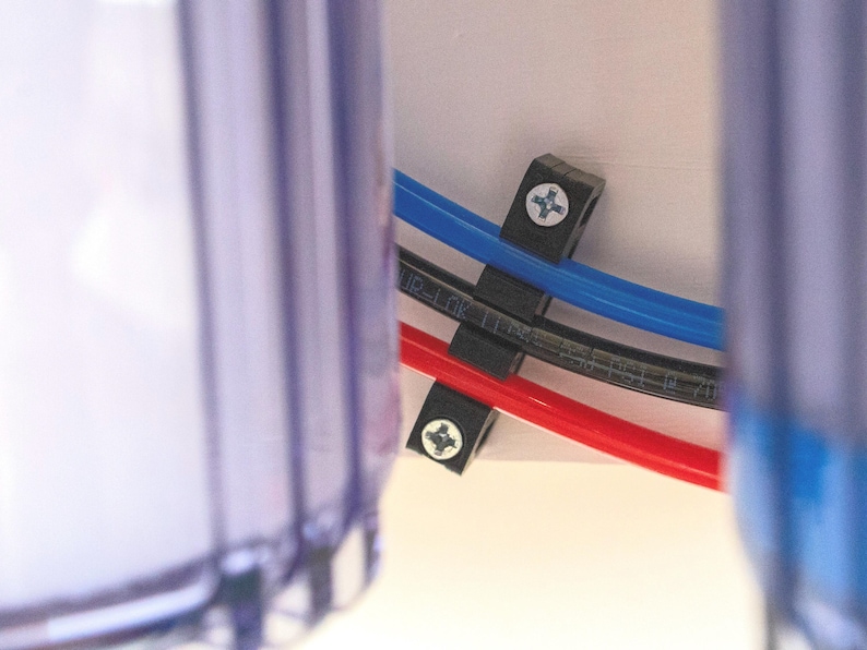 May include: Close-up of three black plastic hose clamps securing three different colored hoses to a white surface. The hoses are red, blue, and black.