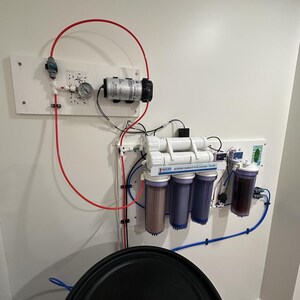 May include: A white wall mounted reverse osmosis water filtration system with six filters, a pressure gauge, and red and blue tubing. The system is connected to a large black container.