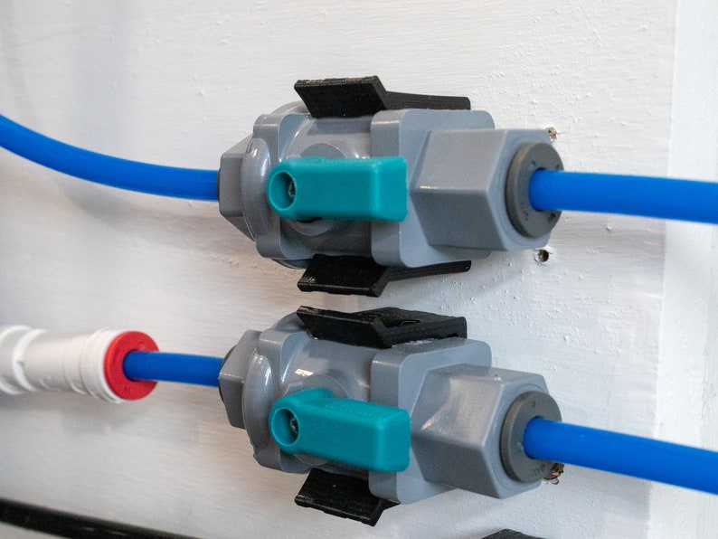 May include: Two gray plastic water valves with blue and teal handles are mounted on a white wall. Each valve has a blue hose attached to it.