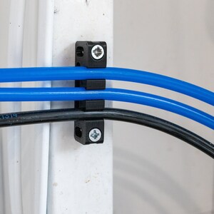 May include: Three blue cables and one black cable are secured to a white wall using a black plastic cable clamp with two screws.