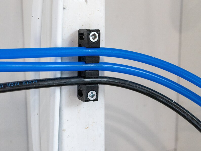May include: Three blue cables and one black cable are secured to a white wall using a black plastic cable clamp with two screws.