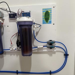 May include: A white wall with a water filtration system, including two clear plastic filters, blue tubing, and two gray valves. There are two digital gauges and a leaf-shaped sticker on the wall.
