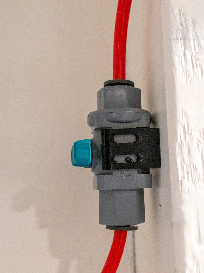 May include: A gray plastic water filter with a blue lever and a red hose attached to it. The filter is mounted on a white wall.