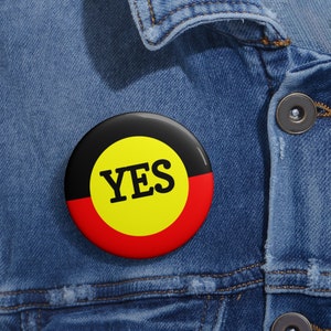 May include: A circular badge featuring a black, yellow, and red design. The word "YES" is printed in bold black letters on a yellow circle. The badge is pinned to a blue denim jacket.