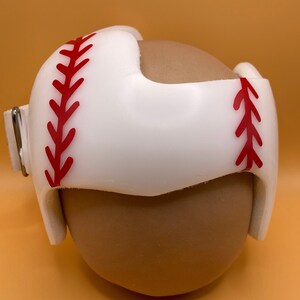 May include: White baseball helmet with red stitching. The helmet has a strap and buckle on the side.