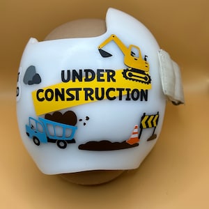 May include: White helmet with a yellow and black construction theme. The helmet has a yellow stripe with the words "UNDER CONSTRUCTION" in black letters. There are images of a yellow excavator, a blue dump truck, a traffic cone, and a construction sign.